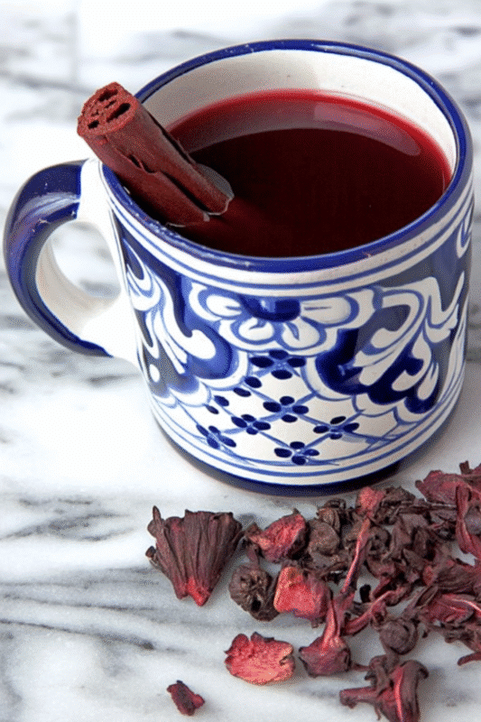 A decorative blue and white mug filled with rich red hibiscus tea, garnished with a cinnamon stick, set against a marble backdrop with scattered dried hibiscus flowers.