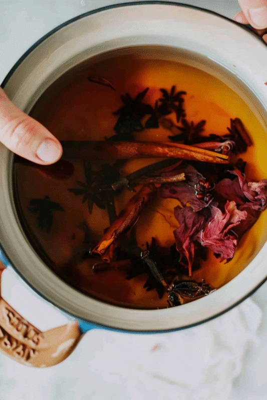 A ceramic mug filled with an amber-colored liquid, garnished with hibiscus flowers, cinnamon sticks, and herbs, creating a cozy and aromatic herbal infusion.