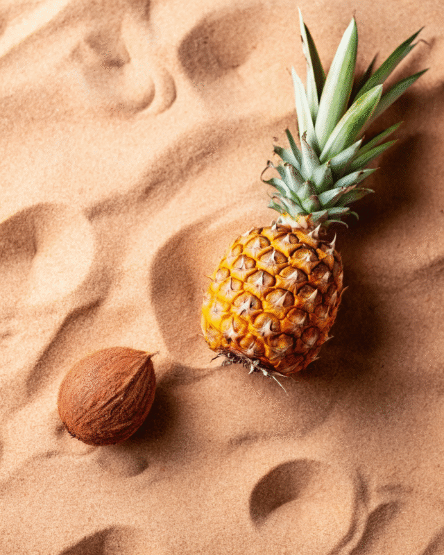 "Fresh pineapple with green leaves and a whole coconut resting on sandy beach, evoking tropical vibes."