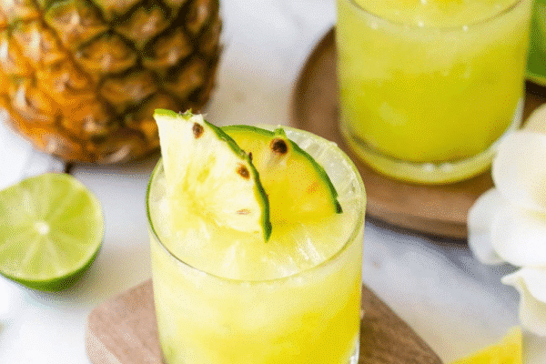"Two glasses of vibrant yellow frozen pineapple coconut water garnished with lime slices, served on wooden coasters with fresh pineapple and flowers in the background."