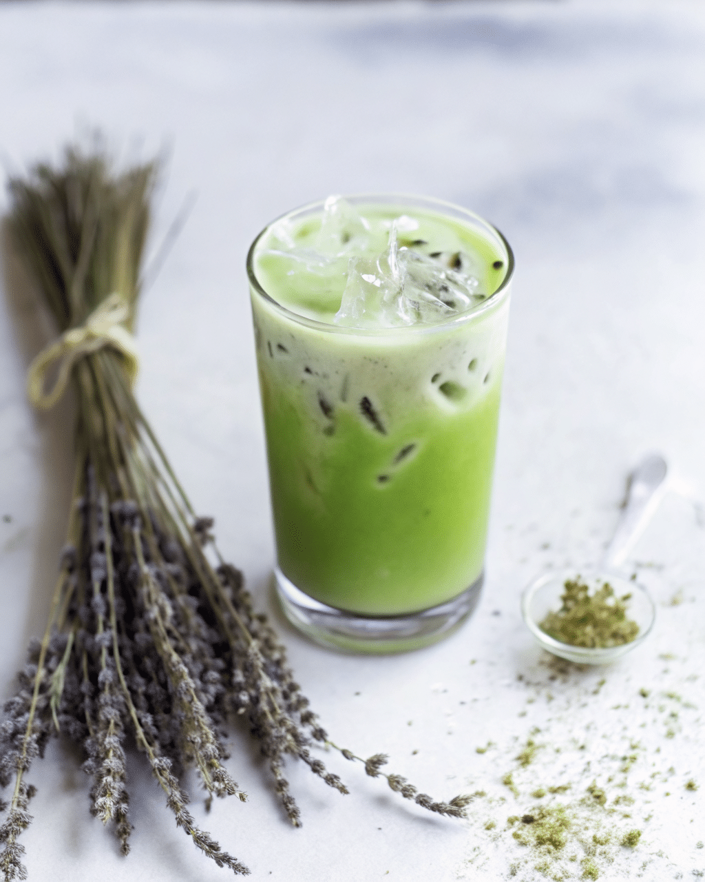 Iced matcha lavender latte in a clear glass with ice cubes, placed on a white surface next to a bundle of dried lavender and a small bowl of matcha powder.