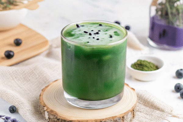 A glass of iced matcha lavender latte topped with black sesame seeds, surrounded by blueberries, dried lavender, and matcha powder on a white surface.