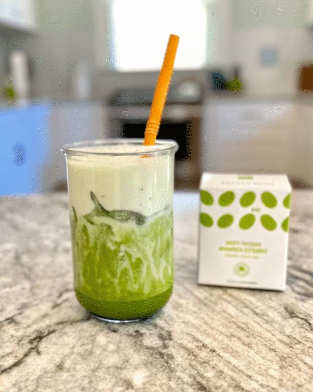 A glass of iced matcha lavender latte with a yellow straw on a marble countertop, with a matcha blend box in the background.