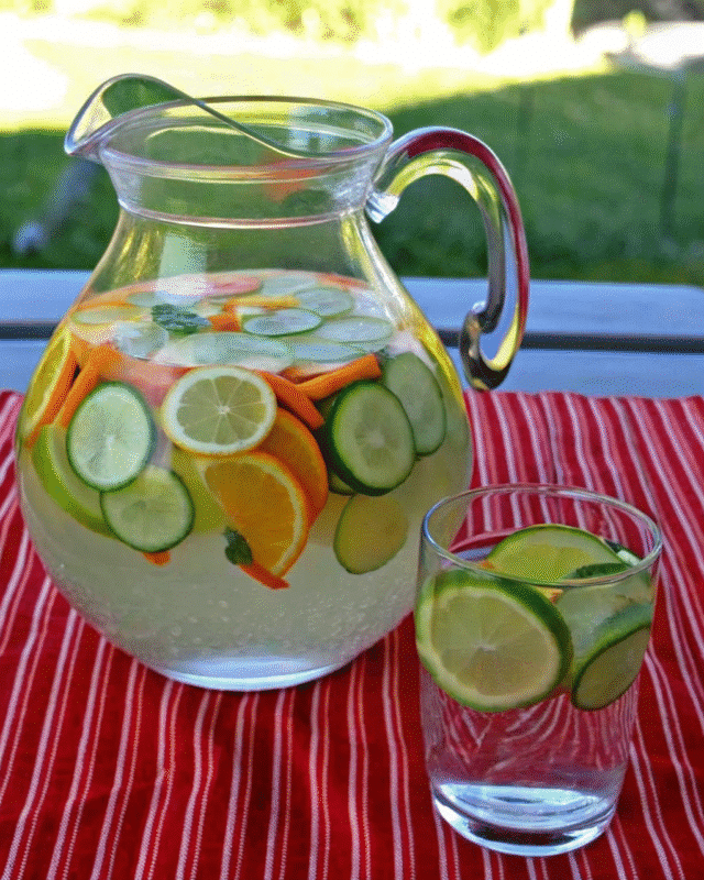 "A refreshing pitcher of infused water filled with slices of cucumber, lemon, lime, orange, and mint, accompanied by a glass garnished with citrus slices on a red-and-white striped tablecloth outdoors."